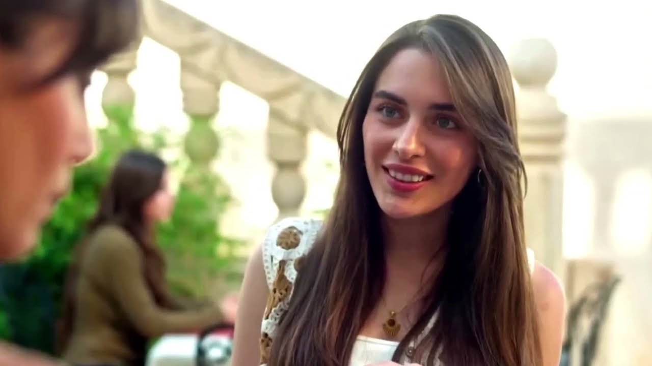 Una giovane donna con i lunghi capelli castano chiaro e un abito bianco ricamato sorride guardando verso l'uomo seduto di fronte a lei, su una terrazza con una balaustra di pietra chiara