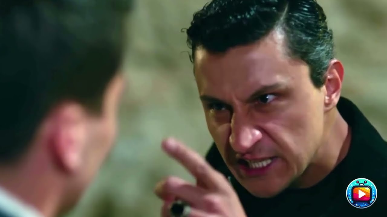 Un uomo in dolcevita nero, con i capelli scuri pettinati all indietro, digrigna i denti e punta l indice contro un altro uomo ripreso di spalle e sfocato in primo piano